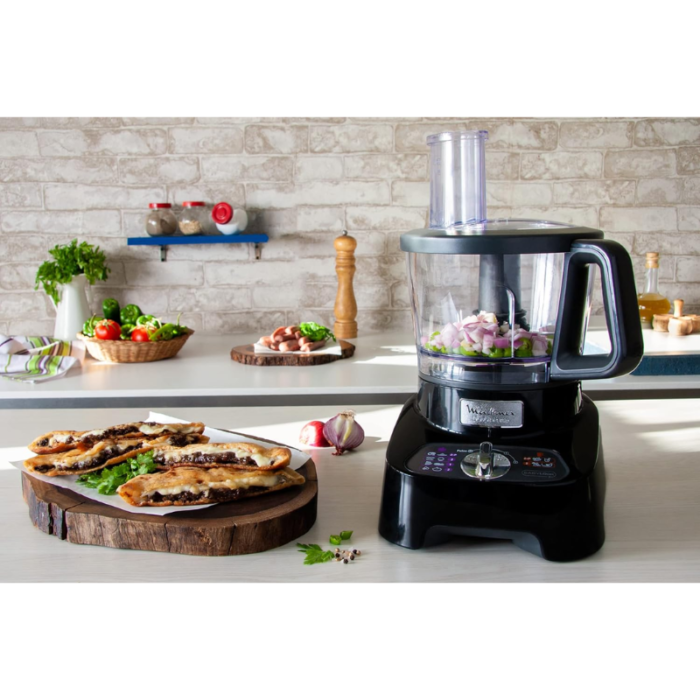A food processor with chopped onions and green peppers inside, placed on a kitchen counter. Fresh vegetables and ingredients are in the background, and cooked flatbread on a wooden board is in the foreground.